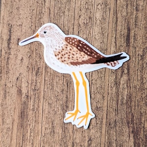 May include: A sticker of a brown and white shorebird with yellow legs and a long, thin beak. The bird is standing on a wooden surface.