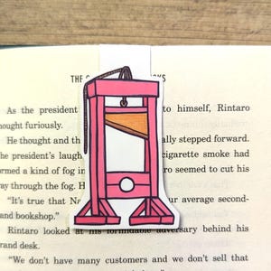 May include: A pink and orange guillotine bookmark. The bookmark is a cartoon illustration of a guillotine with a rope and a blade. The bookmark is placed on a page of a book with text.