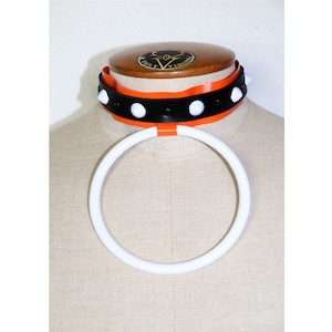 May include: A black leather choker with white studs and an orange trim. The choker is on a white mannequin. A white plastic ring is around the mannequin's neck.