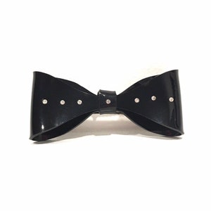 Könnte beinhalten: Eine schwarze Schleife mit Strasssteinen. Die Schleife ist aus glänzendem Material und hat ein schlichtes Design.