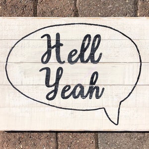 May include: White wooden sign with black lettering in a speech bubble that reads "Hell Yeah".