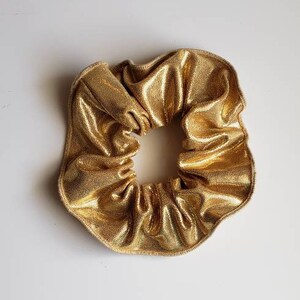 May include: A gold scrunchie made of shiny fabric. The scrunchie is gathered in a circle and has a smooth, even texture.