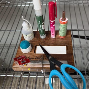 May include: A wooden sewing organizer with the text Sew Happy Sewin! engraved on the front. It holds a small glue bottle, an Easy Press Pen, a pink marker, a spool of green thread, a blue pincushion, two red clips, sewing pins, and blue scissors.