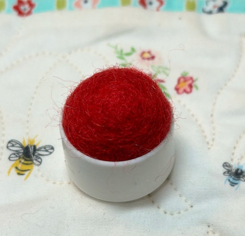 May include: A red pincushion in a white ceramic dish. The pincushion is made of wool and is very fluffy.
