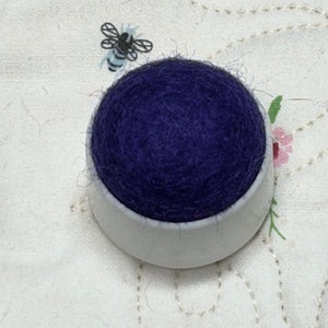 May include: A purple pincushion in a white ceramic base. The pincushion is made of a soft, fluffy material.