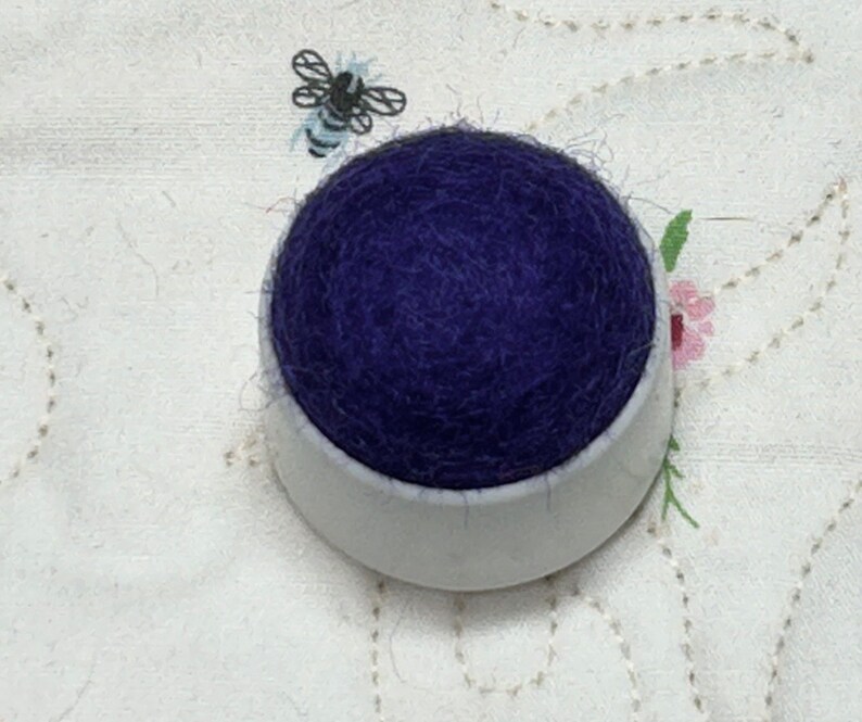 May include: A purple pincushion in a white ceramic base. The pincushion is made of a soft, fluffy material.