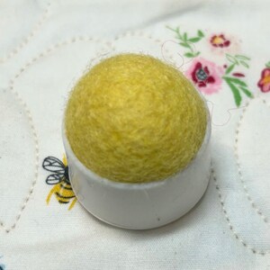 May include: A yellow wool pincushion sits on a white plastic base. The pincushion is round and has a fuzzy texture.
