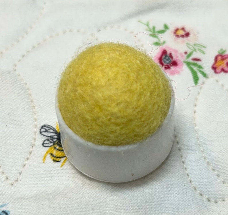 May include: A yellow wool pincushion sits on a white plastic base. The pincushion is round and has a fuzzy texture.