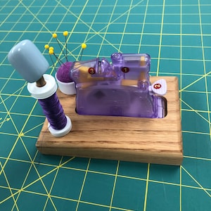 May include: A wooden sewing thread organizer with a purple thread spool, a purple pincushion with yellow pins, and a clear purple thread cutter with the numbers 1, 2, and 3.