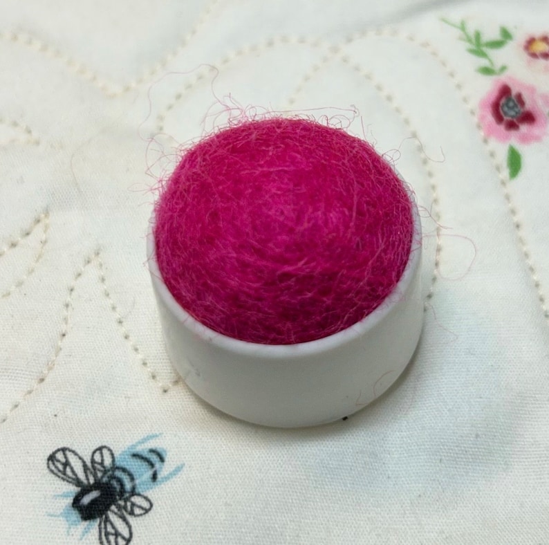 May include: A pink wool pincushion in a white plastic container.