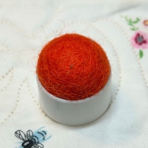 May include: A bright orange pincushion with a white plastic base. The pincushion is made of wool and is round.