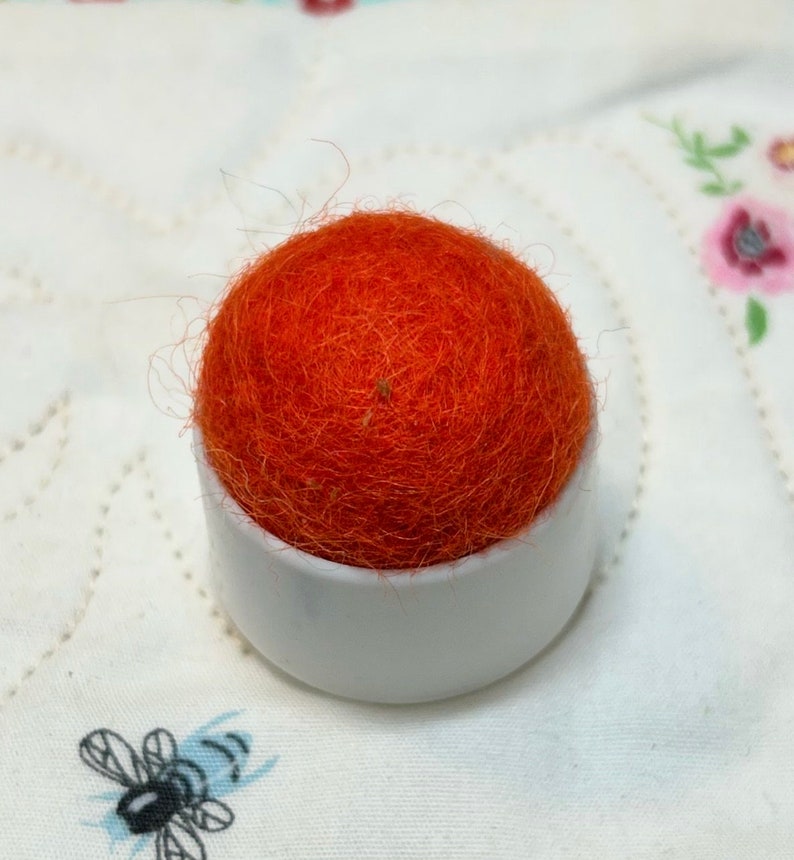 May include: A bright orange pincushion with a white plastic base. The pincushion is made of wool and is round.