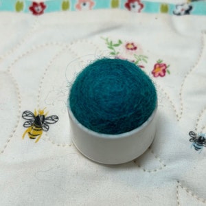 May include: A teal felt pincushion sits on a white plastic base. The pincushion is round and has a textured surface.