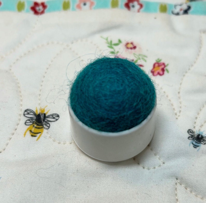 May include: A teal felt pincushion sits on a white plastic base. The pincushion is round and has a textured surface.