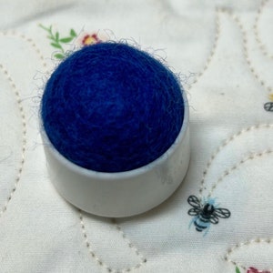 May include: A blue felt pincushion in a white plastic base. The pincushion is sitting on a white fabric with a floral pattern.