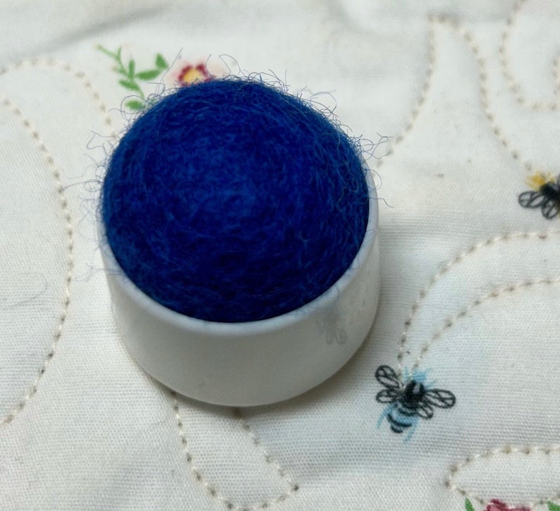 May include: A blue felt pincushion in a white plastic base. The pincushion is sitting on a white fabric with a floral pattern.