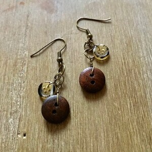 May include: A pair of handmade earrings with dark brown wooden buttons and small, translucent buttons. The earrings have bronze-coloured hooks and connecting chains. The wooden buttons are round with two holes, and the smaller buttons have a patterned design.