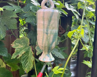 Puede incluir: Un comedero para colibríes de cerámica hecho a mano con una base beige y goteos de esmalte verde y azul. El comedero tiene una punta de plástico roja y está suspendido por una cadena de metal. El fondo presenta follaje verde.