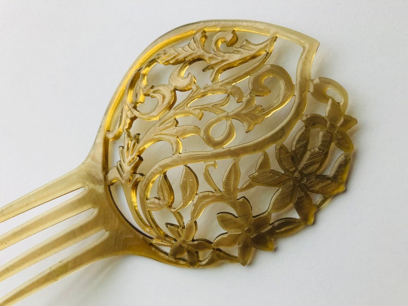 May include: A vintage-style hair comb with intricate floral and leaf designs. The comb is a light amber color with a detailed, openwork pattern. The design features flowers and swirling leaves, creating an elegant accessory.