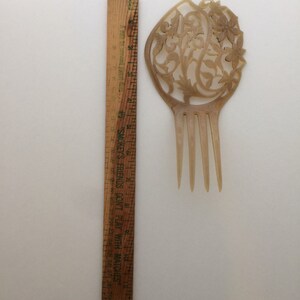 May include: A vintage, light brown hair comb with an ornate floral design. The comb is approximately 4 inches long and features a decorative top with carved flowers and vines. A wooden ruler is next to the comb for scale.