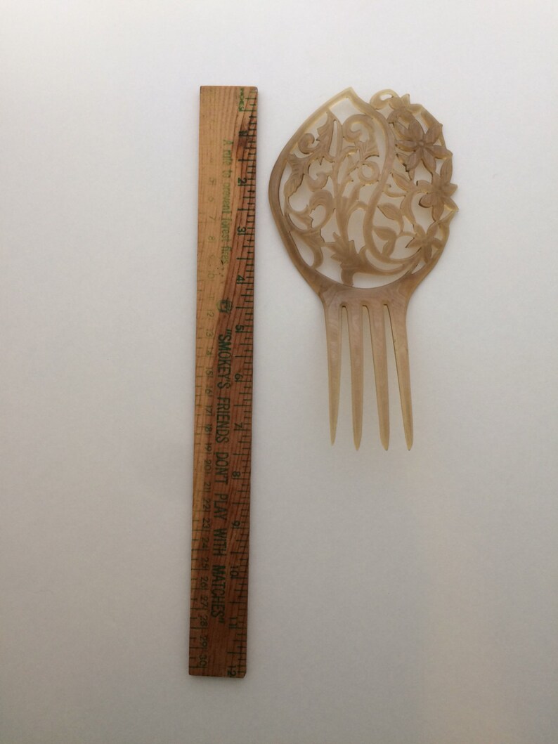 May include: A vintage, light brown hair comb with an ornate floral design. The comb is approximately 4 inches long and features a decorative top with carved flowers and vines. A wooden ruler is next to the comb for scale.