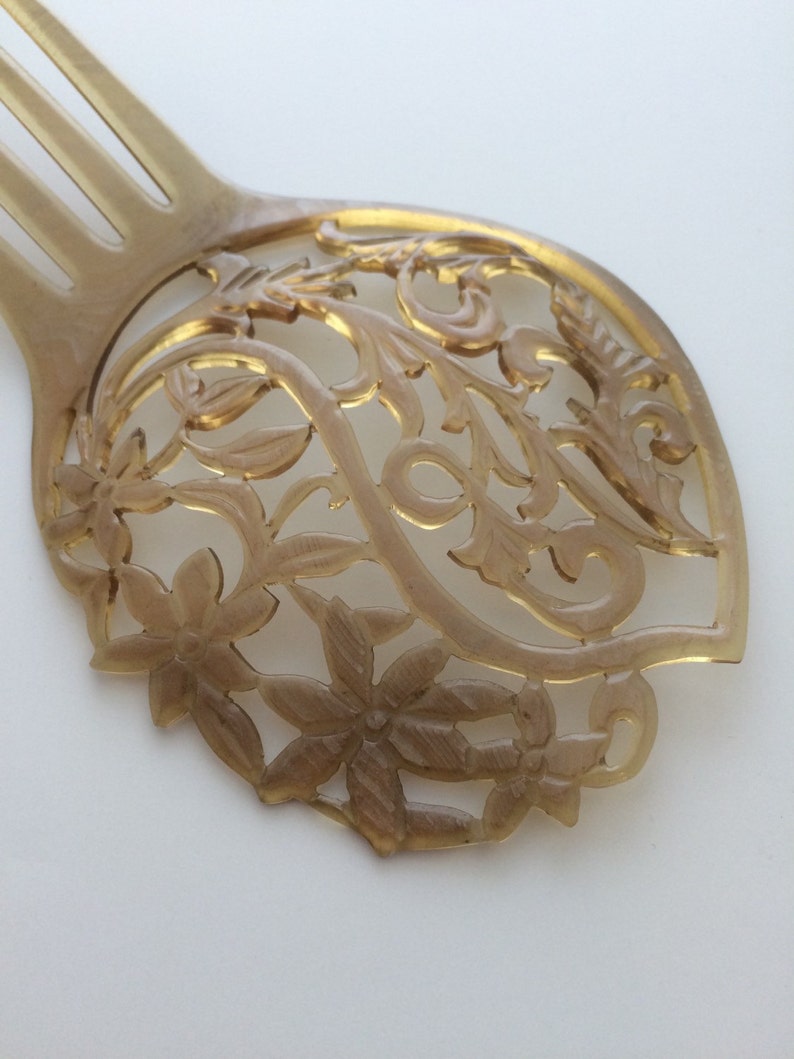 May include: A light brown hair comb with a decorative floral design. The comb features a series of tines and a detailed, openwork pattern of flowers and leaves. The comb is made of a translucent material.
