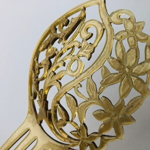 May include: A golden-colored hair comb with an intricate floral and leaf design. The comb features a decorative openwork pattern with detailed flowers and foliage. The tines of the comb are visible at the base, and the overall design is elegant.