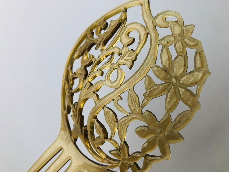 May include: A golden-colored hair comb with an intricate floral and leaf design. The comb features a decorative openwork pattern with detailed flowers and foliage. The tines of the comb are visible at the base, and the overall design is elegant.