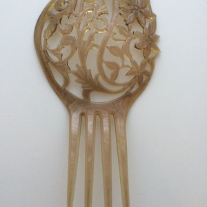 May include: A vintage hair comb with a decorative floral design. The comb is made of a light brown material with intricate cut-out patterns and four long teeth. The top features a floral and leaf motif.
