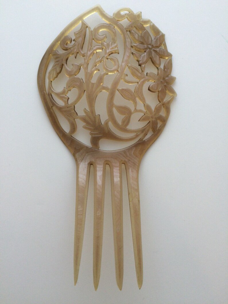 May include: A vintage hair comb with a decorative floral design. The comb is made of a light brown material with intricate cut-out patterns and four long teeth. The top features a floral and leaf motif.