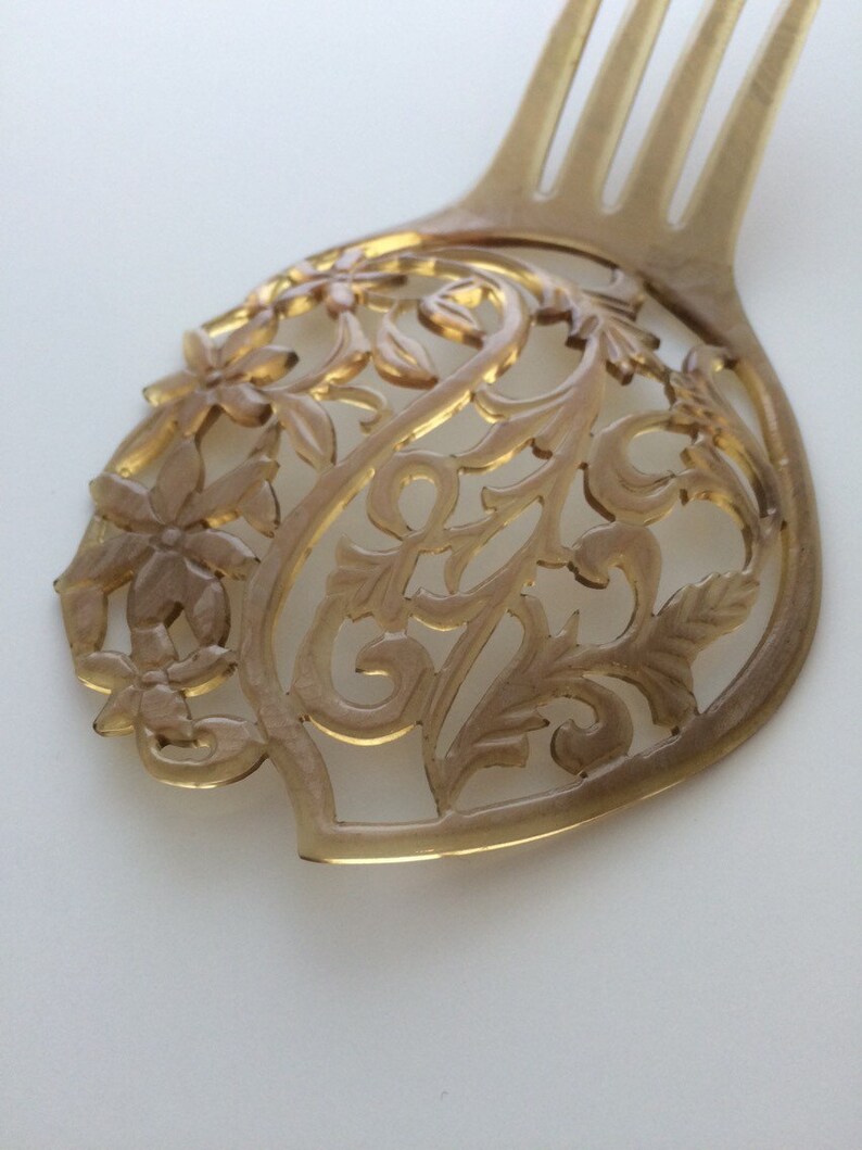 May include: A light brown hair comb with a decorative floral design. The comb features a detailed, openwork pattern of flowers and leaves. The tines of the comb are visible at the top.