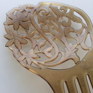 May include: A vintage-style hair comb in a light amber color. The comb features an intricate floral and leaf design at the top, with a series of teeth extending downwards. The design is carved out, creating an openwork effect.