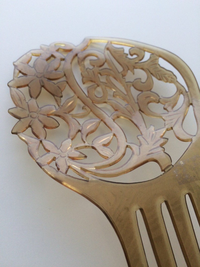 May include: A vintage-style hair comb in a light amber color. The comb features an intricate floral and leaf design at the top, with a series of teeth extending downwards. The design is carved out, creating an openwork effect.