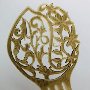 May include: A light brown hair comb with an intricate floral and leaf design. The comb features a decorative top with carved flowers and swirling patterns. The tines of the comb are visible at the bottom.