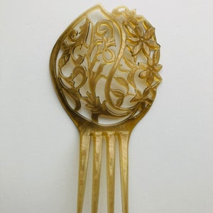 May include: A vintage hair comb with a floral and leaf design. The comb is a light amber color and features intricate cut-out details. The comb has four tines and a decorative top with flowers and swirling patterns.