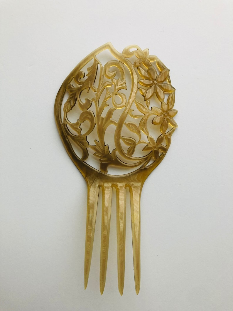 May include: A vintage hair comb with a floral and leaf design. The comb is a light amber color and features intricate cut-out details. The comb has four tines and a decorative top with flowers and swirling patterns.