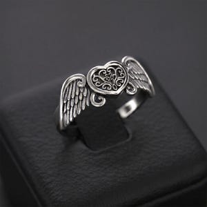Pu&ograve; includere: Un anello color argento caratterizzato da un elemento centrale a forma di cuore con intricati motivi a filigrana. Il cuore &egrave; affiancato da due ali dettagliate e testurizzate che si estendono verso l'esterno lungo la fascia dell'anello.