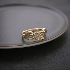 Pu&ograve; includere: Un anello color oro caratterizzato da un elemento centrale a forma di cuore con un intricato motivo in filigrana. Il cuore &egrave; affiancato da due ali dettagliate e strutturate che si estendono lungo la fascia, creando un design coeso.