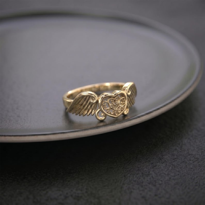 Pu&ograve; includere: Un anello color oro caratterizzato da un elemento centrale a forma di cuore con un intricato motivo in filigrana. Il cuore &egrave; affiancato da due ali dettagliate e strutturate che si estendono lungo la fascia, creando un design coeso.