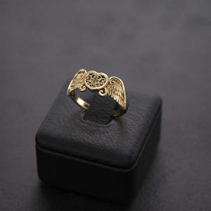 Pu&ograve; includere: Un anello color oro caratterizzato da un motivo centrale a forma di cuore con intricati dettagli in filigrana, affiancato da due ali piumate stilizzate che si estendono dai lati. La superficie metallica presenta una finitura lucida.