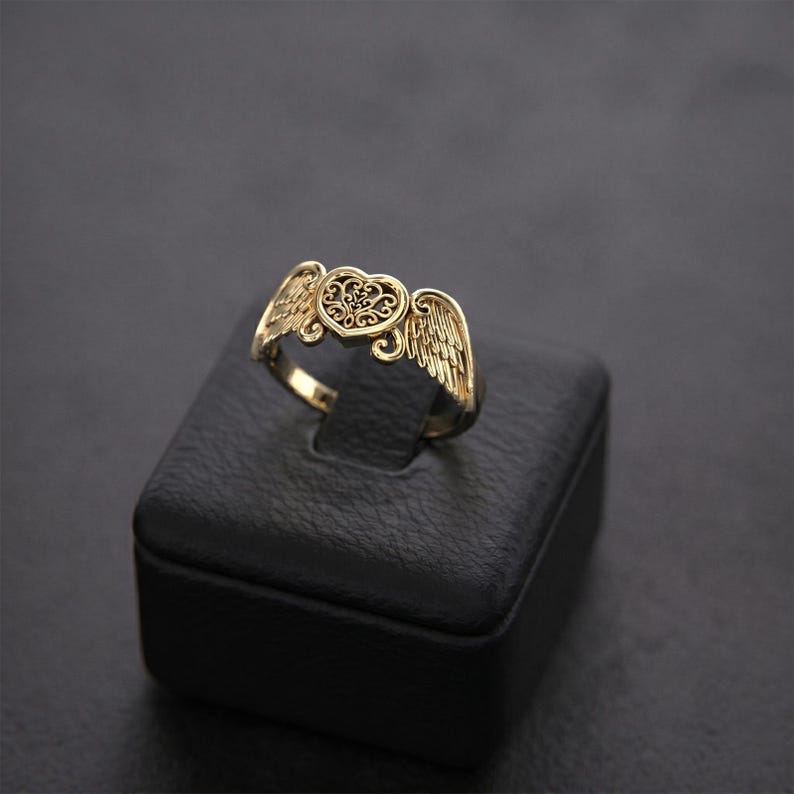 Pu&ograve; includere: Un anello color oro caratterizzato da un motivo centrale a forma di cuore con intricati dettagli in filigrana, affiancato da due ali piumate stilizzate che si estendono dai lati. La superficie metallica presenta una finitura lucida.
