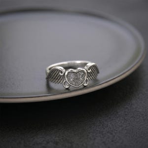 Pu&ograve; includere: Un anello in metallo color argento con un elemento centrale a forma di cuore caratterizzato da un intricato motivo a filigrana. Il cuore &egrave; affiancato da due ali dettagliate che si estendono verso l'esterno, creando un design simmetrico.