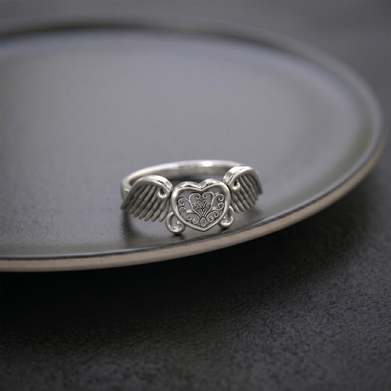 Pu&ograve; includere: Un anello in metallo color argento con un elemento centrale a forma di cuore caratterizzato da un intricato motivo a filigrana. Il cuore &egrave; affiancato da due ali dettagliate che si estendono verso l'esterno, creando un design simmetrico.