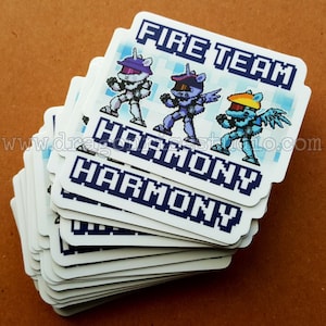 May include: A stack of white stickers with a blue pixelated design. The stickers feature three pixelated characters in blue, purple, and yellow, with the text "FIRE TEAM" and "HARMONY" in blue.