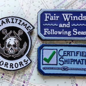 Op de afbeelding: Drie geborduurde patches met nautische thema's. De eerste patch is zwart-wit met een schedel en gekruiste beenderen en de tekst "Maritime Horrors". De tweede patch is blauw met witte tekst die "Fair Winds and Following Seas" luidt. De derde patch is blauw met een groen vinkje en de tekst "Certified Shipmate".