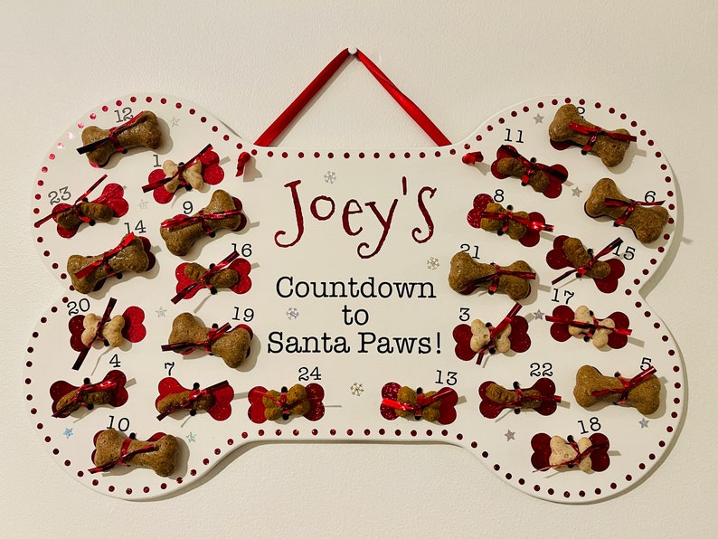 May include: A white bone-shaped wooden advent calendar with red bows and dog treats. The calendar is decorated with red dots and silver snowflakes. The text "Joey's Countdown to Santa Paws!" is printed on the calendar.
