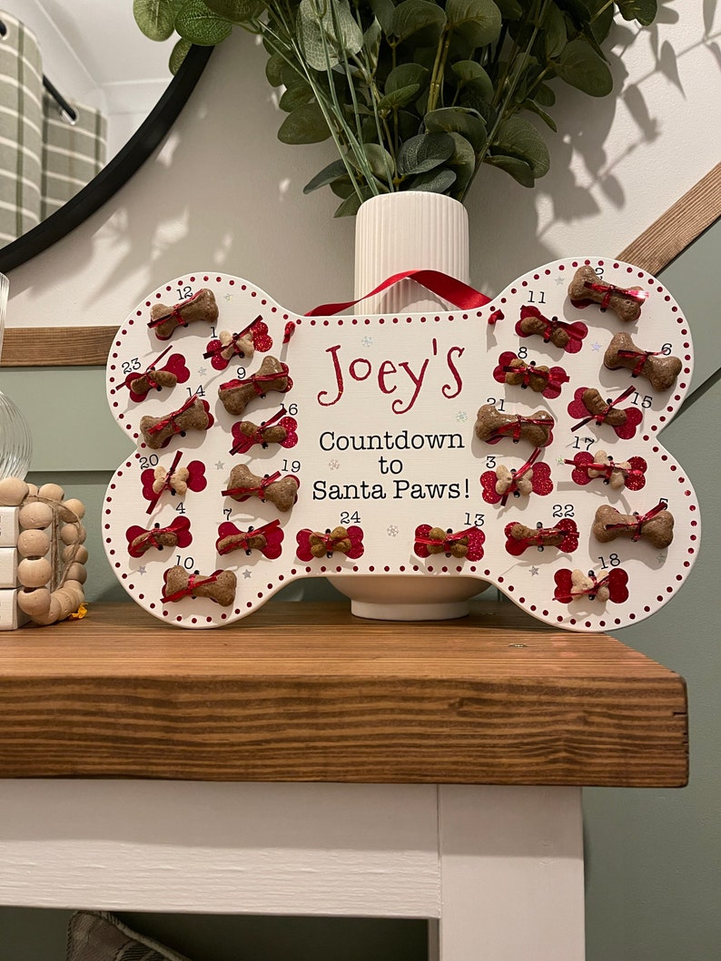 May include: White bone-shaped advent calendar with red bows and dog treats. The calendar is decorated with red dots and snowflakes. The text "Joey's Countdown to Santa Paws!" is written on the calendar.