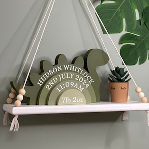 May include: Green wooden dinosaur-shaped baby birth announcement with the text "Hudson Whitlock 2nd July 2024 11:09AM 7lb 2oz" hanging from a white rope on a white shelf.
