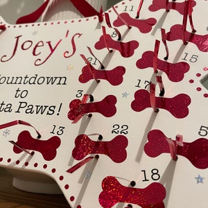 May include: White wooden dog bone shaped advent calendar with red glitter bone cutouts. Each bone has a number from 1 to 22. The calendar is titled "Joey's Countdown to Santa Paws!"
