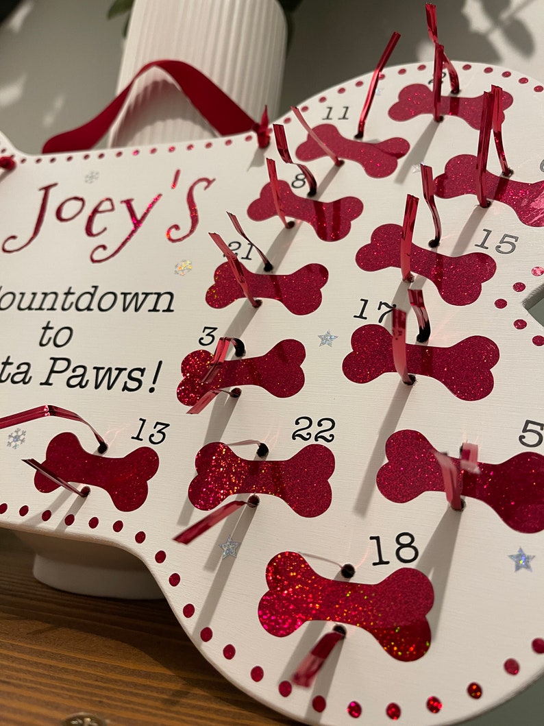 May include: White wooden dog bone shaped advent calendar with red glitter bone cutouts. Each bone has a number from 1 to 22. The calendar is titled "Joey's Countdown to Santa Paws!"